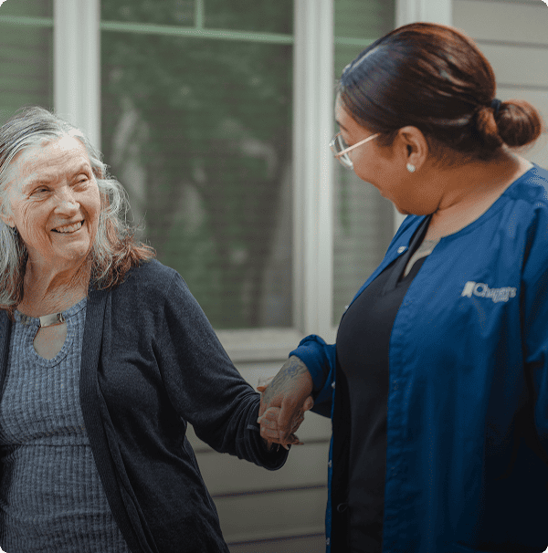 An elderly woman smiles while holding hands with a caregiver in blue scrubs outside a house, both appearing happy and engaged in conversation. An elderly woman smiles while holding hands with a caregiver in blue scrubs outside a house, both appearing happy and engaged in conversation.